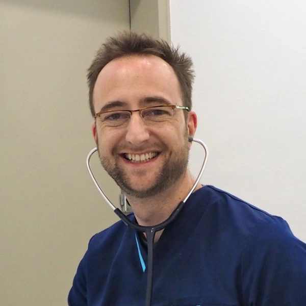 Docteur Vétérinaire Hugues Journaux souriant en blouse bleue portant un stéthoscope autour du cou.